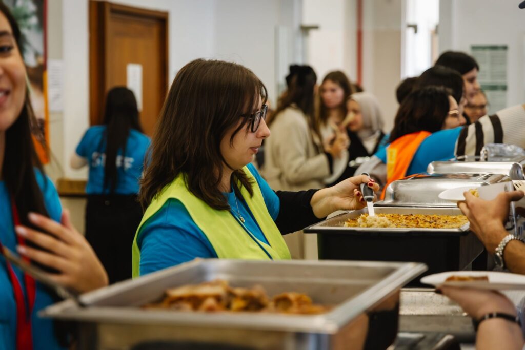 Voluntaria sirviendo comida en un comedor comunitario, usando chaleco reflectante amarillo y camiseta azul, con varias personas en fila esperando su turno.