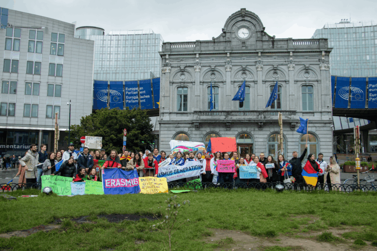 a group of international students and ESN volunteers standing in Luxembourg Square with banners and flags for a group picture. On the back there’s the european parliament.