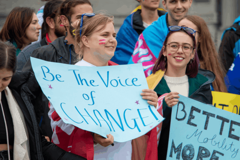 Youth activists advocating for better student mobility and change, smiling and holding colorful protest signs “Be the voice of CHANGE” at an Erasmus Generation parade in Brussels