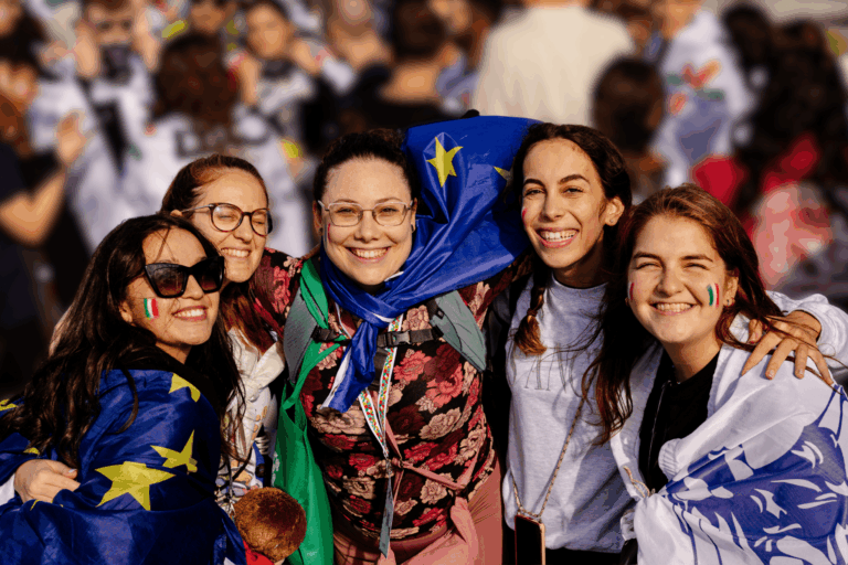 Grupo de estudiantes Erasmus sonriendo y abrazándose durante un evento internacional. Llevan banderas y pinturas faciales que representan diferentes países, destacando la amistad, inclusión y diversidad cultural del programa Erasmus.
