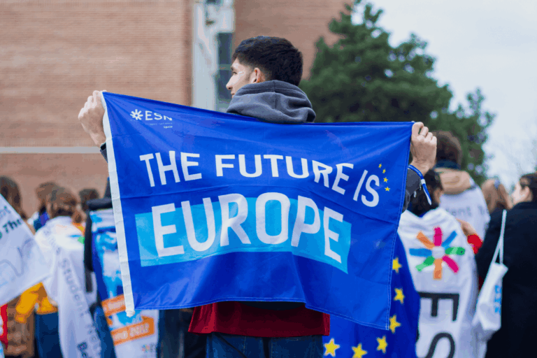 Joven sostiene una bandera azul con el mensaje “The future is Europe” durante una actividad de Erasmus Student Network junto a otros voluntarios.