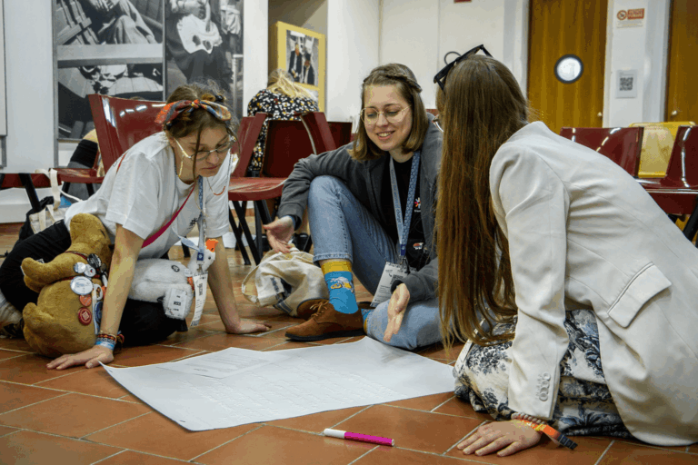 Tres jóvenes adultas participan activamente en una actividad grupal en el suelo, revisando un póster con ideas escritas durante un taller Erasmus+.