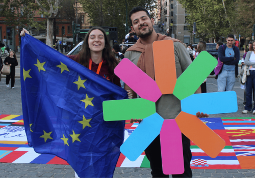 web12 Dos jóvenes sonrientes posan al aire libre durante un evento Erasmus. Ella sostiene una bandera de la Unión Europea y él un símbolo colorido en forma de estrella, representando a ESN. Están de pie sobre una gran alfombra con banderas de distintos países. A su alrededor, más personas participan en la actividad en una plaza urbana rodeada de árboles y edificios históricos.