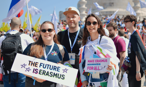 web2 Tres jóvenes sonrientes posan frente a un monumento, rodeados de una multitud con banderas y carteles. Dos de ellos sostienen letreros: uno dice “THE FUTURE IS ERASMUS” y el otro “MAKE ERASMUS accessible FOR ALL”. Visten camisetas y credenciales del programa Erasmus, y una de las chicas lleva varias banderas europeas sobre los hombros. El ambiente es festivo y soleado.