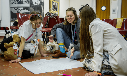 web9 Tres jóvenes adultas participan activamente en una actividad grupal en el suelo, revisando un póster con ideas escritas durante un taller Erasmus+.